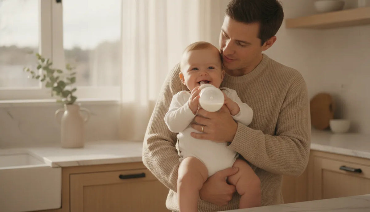 A parent gently feeding a smiling baby a bottle in a bright, minimal kitchen