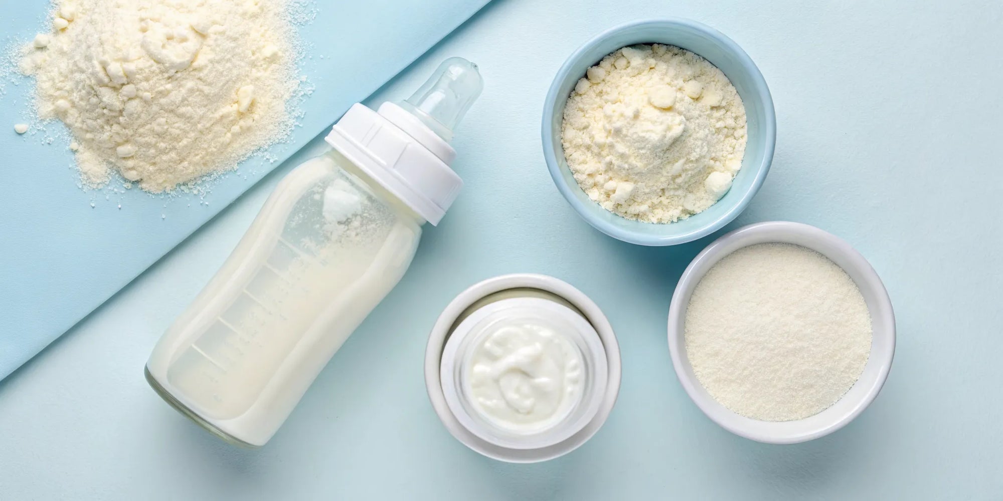 A baby bottle surrounded by bowls of different baby formula ingredients.